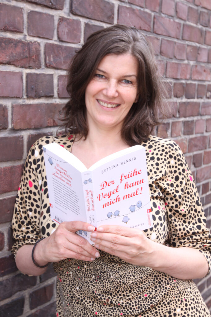 Die Autorin Bettina Hennig steht an einer Ziegelsteinmauer und hält ein Buch in der Hand. Sie hat halblanges braunes Haar und lächelt in die Kamera.