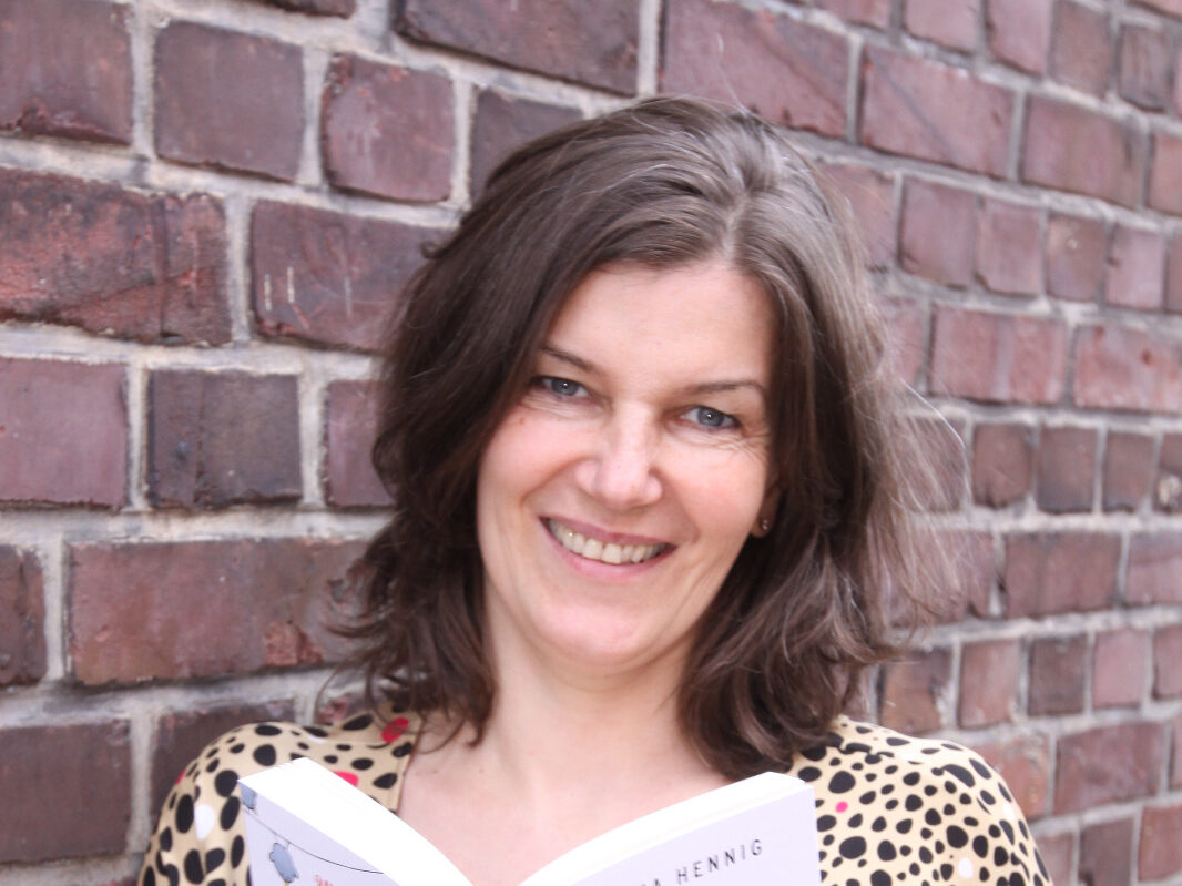 Die Autorin Bettina Hennig steht an einer Ziegelsteinmauer und hält ein Buch in der Hand. Sie hat halblanges braunes Haar und lächelt in die Kamera.